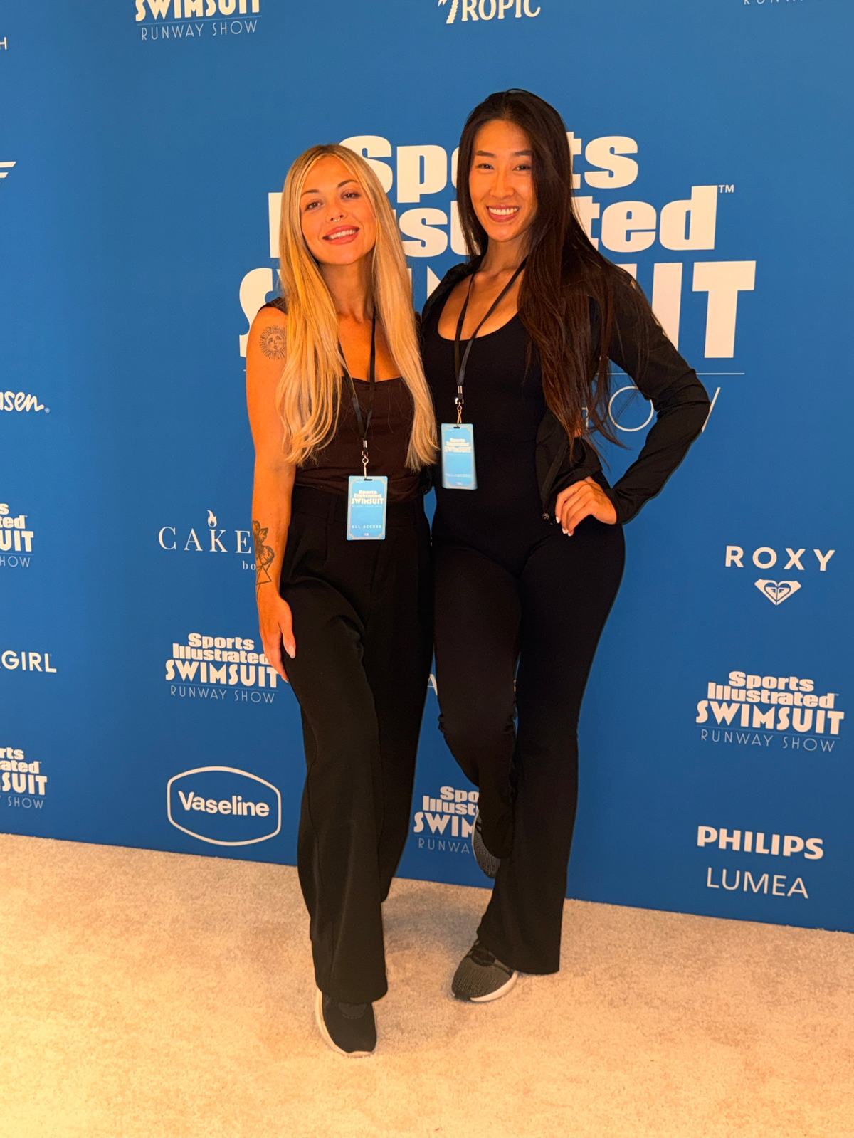 Alivia with a crew member in front of the Sports Illustrated Swimsuit Runway Show step-and-repeat, wearing all-access badges